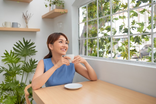 녹색 창 뒤에 커피를 마시는 아름다운 아시아 여자