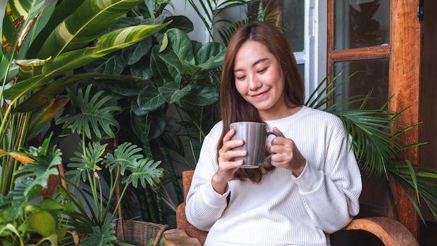 ホットコーヒーを飲み、自宅の観葉植物の庭のあるバルコニーでリラックスした美しいアジアの女性