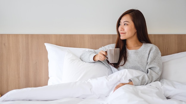 A beautiful asian woman drinking hot coffee while lying on a white cozy bed at home in the morning