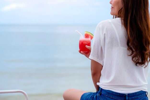 海のそばに座ってスイカジュースを飲む美しいアジアの女性