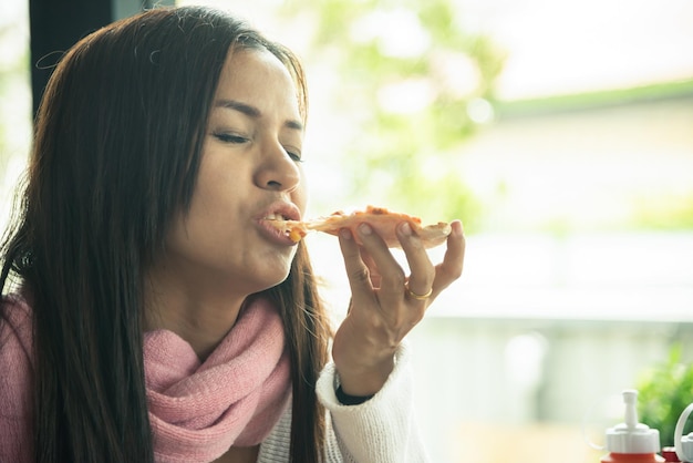 ピザのスライスを食べる美しいアジアの女性。レストランでピザを食べるセーターの面白いアジアの女の子。