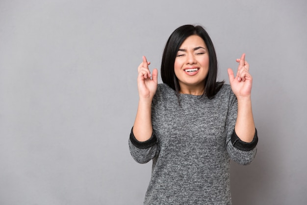 Beautiful asian woman in gray jumper keeping fingers crossed and eyes closed