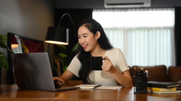 Bella donna asiatica che tiene tazza di caffè e utilizza il computer portatile sullo scrittorio di legno