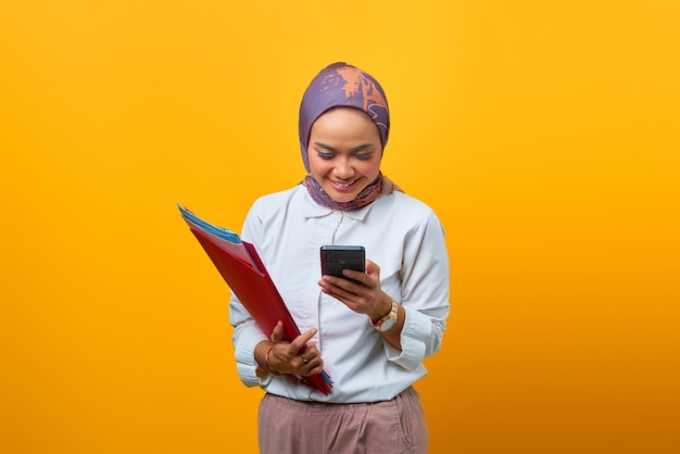 Beautiful Asian woman holding folders and using smartphone over yellow background