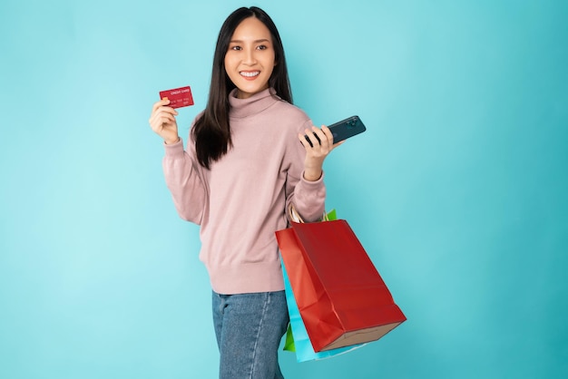 Foto bella donna asiatica che tiene borse della spesa di carta e mostra la carta di credito con lo smartphone su sfondo blu