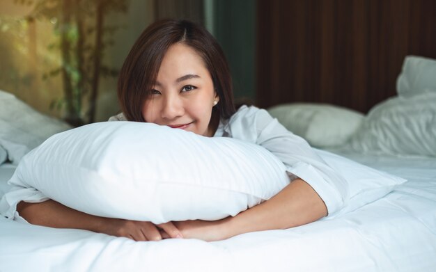 Foto una bella donna asiatica in possesso di un cuscino mentre sdraiato su un letto accogliente bianco a casa