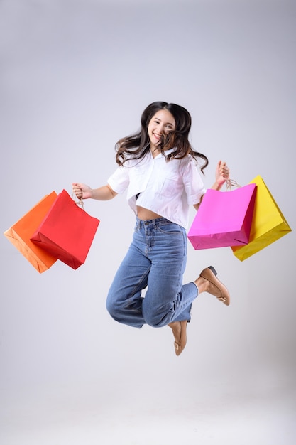 Foto bella donna asiatica tenendo le borse della spesa di vari colori e saltando con una felice espressione su uno sfondo bianco.