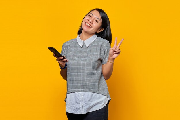 Beautiful Asian woman holding smartphone smiling and showing peace sign over yellow background