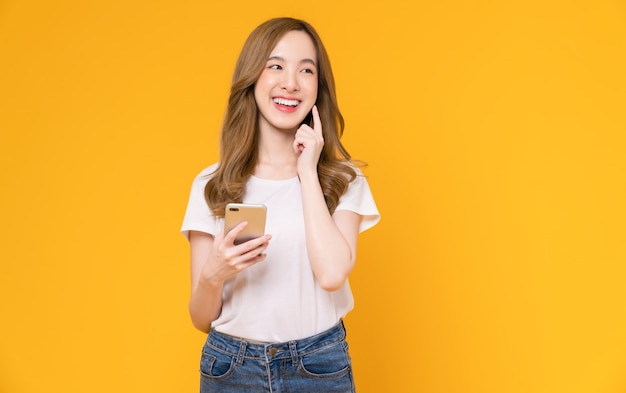 Beautiful Asian woman holding smartphone and smiling with typing on mobile, light yellow background.