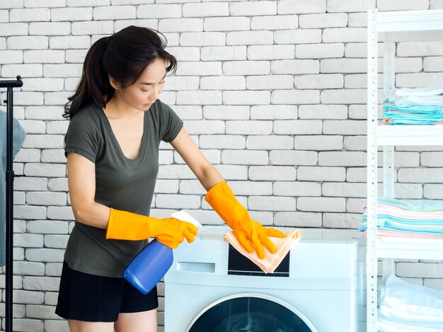 Beautiful Asian woman, housewife wearing orange protective rubber gloves using cleaner spray and clean cloth to cleaning washing machine on white brick wall