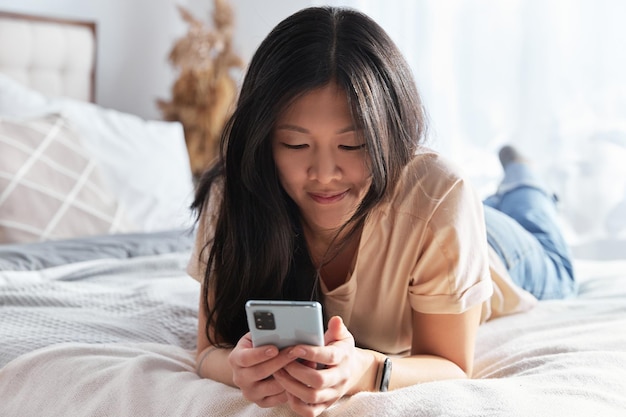 사진 침대에 누워있는 스마트폰을 사용하는 캐주얼 옷을 입은 아름다운 아시아 여성