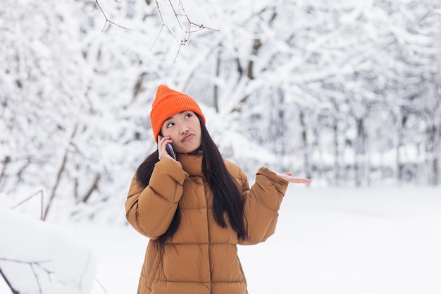 散歩の冬の雪の日に公園で電話で話している暖かい服を着た美しいアジアの女性