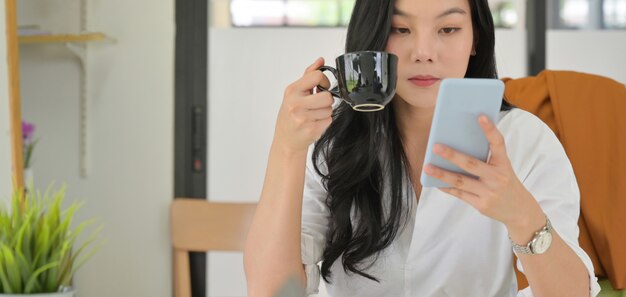 La bella donna asiatica sta controllando i messaggi su uno smartphone e sta bevendo il caffè in un ufficio comodo.