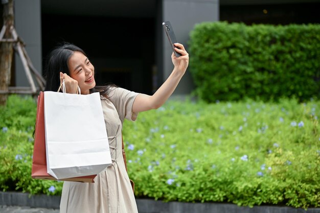 A beautiful Asian woman is taking selfies with her shopping bags while walking on the street