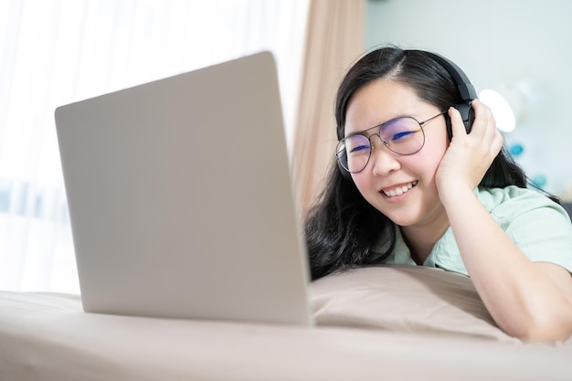 아름다운 아시아 여성이 노트북이 있는 헤드셋을 사용하고 파스텔 녹색 갈색 테마로 침실에 있는 침대에 누워 있습니다.