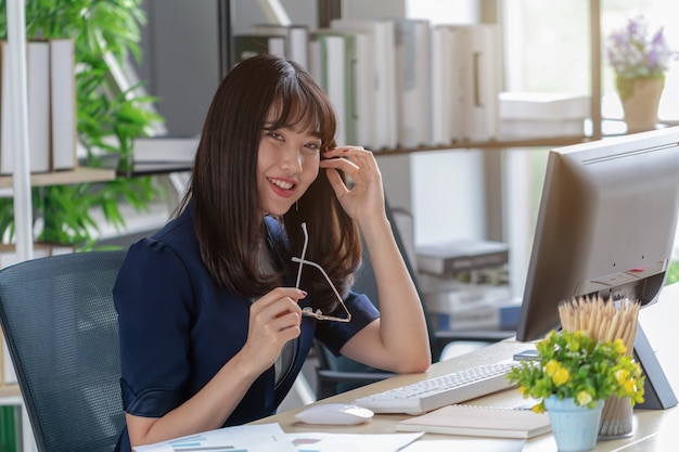 A beautiful Asian woman is wearing a dark blue suit sitting at a desk in a modern office and is enjoying her work.