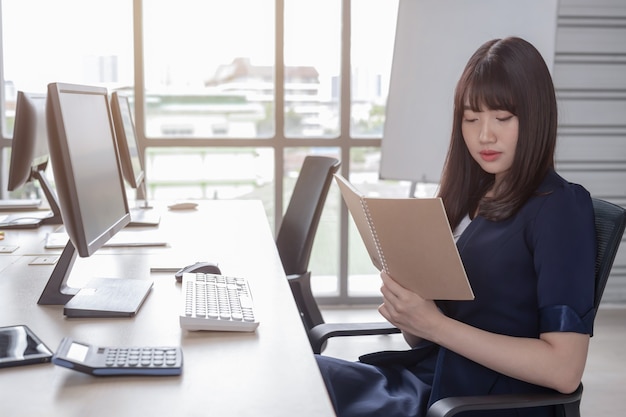A beautiful Asian woman is wearing a dark blue suit sitting at a desk in a modern office and is enjoying her work.
