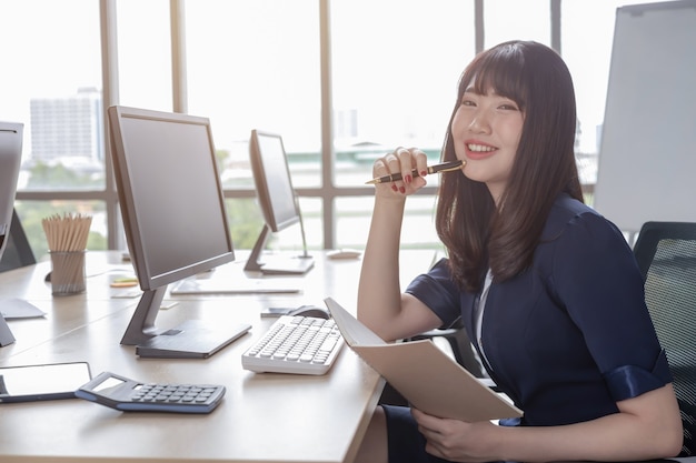 Una bella donna asiatica indossa un abito blu scuro seduto a una scrivania in un ufficio moderno ed è felice di lavorare e ha uno sfondo di una grande finestra di vetro.