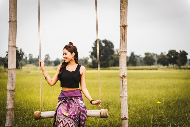 Photo beautiful asian woman in local dress sitting on swing and enjoy natural on bamboo bridge in rice field