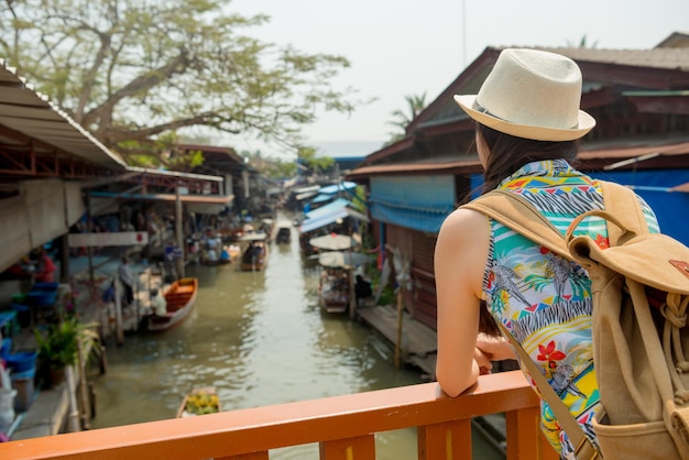 beautiful asian woman looking at Damnoen Saduak floating market standing on the land and watching all pedlar shop lifestyle in the Thailand during travel vacation in summer.