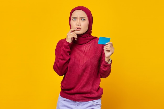 Beautiful Asian woman looking stressed and nervous with biting nails and showing credit card over yellow background