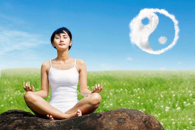 Beautiful Asian woman meditating outdoors on the rock at meadow with yin yang clouds symbol in the sky