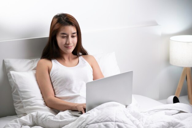 Beautiful Asian woman in pajamas using laptop sitting on the bed in the house