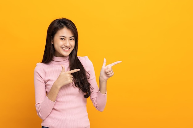 Beautiful asian woman pointing finger into blank space Smiling female wearing pink shirt on isolated yellow background Attractive Teenager girl looking at empty space for text