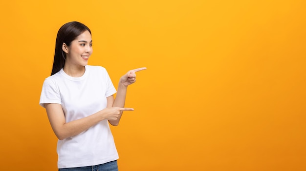 Beautiful asian woman pointing finger into blank space. smiling female wearing white shirt on isolated yellow background. attractive teenager girl looking at empty space for text