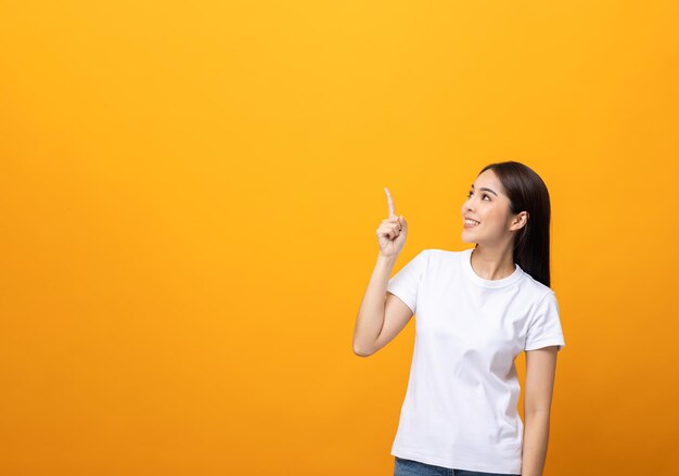 Beautiful asian woman pointing finger into blank space. Smiling female wearing white shirt on isolated yellow background. Attractive Teenager girl looking at empty space for text.