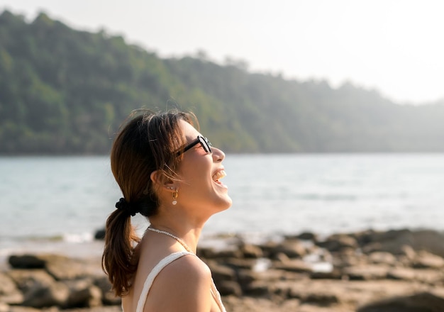 Beautiful Asian woman portrait with sunglasses on the beach in summer vibes Happy female smile with teeth looking up to sky on sea island rocks and sky background Holiday vacation summertime