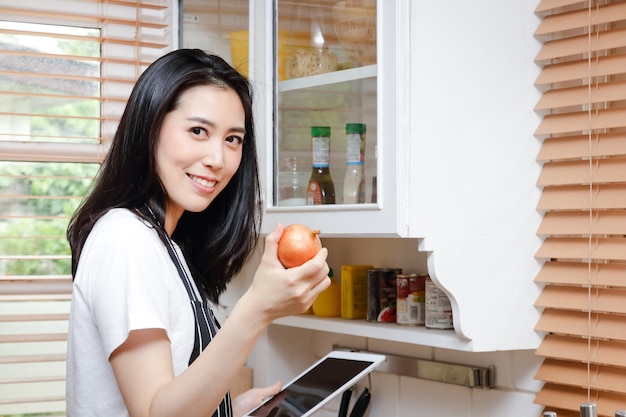 Beautiful asian woman practicing cooking in the kitchen at home\
hold your tablet to watch online cooking videos. concept of living\
during the coronavirus pandemic. social distancing