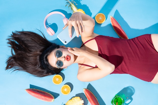 빨간 비키니, 선글라스와 헤드폰, 전화 및 과일 주위에 거짓말에 아름 다운 아시아 여자. 열 대 여름 해변 여행 컨셉