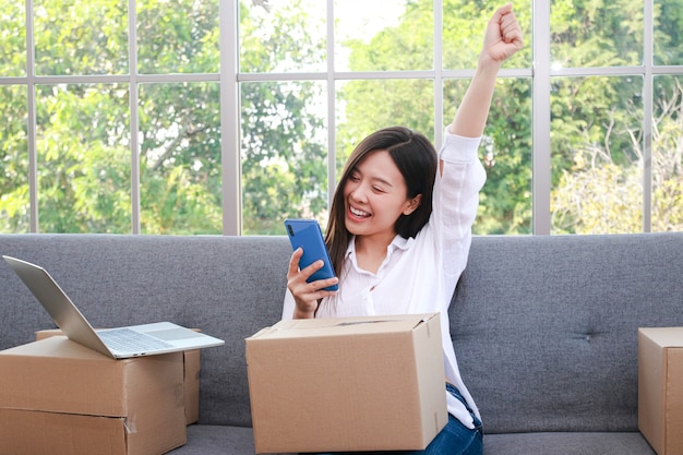 Красивая азиатская женщина продает онлайн Поднимите руку, чтобы радоваться получению заказов на смартфоны. предпринимательская концепция Ведение бизнеса в Интернете, отправка товаров по почте.