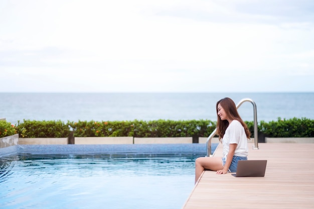 Una bella donna asiatica seduta a bordo piscina con un computer portatile