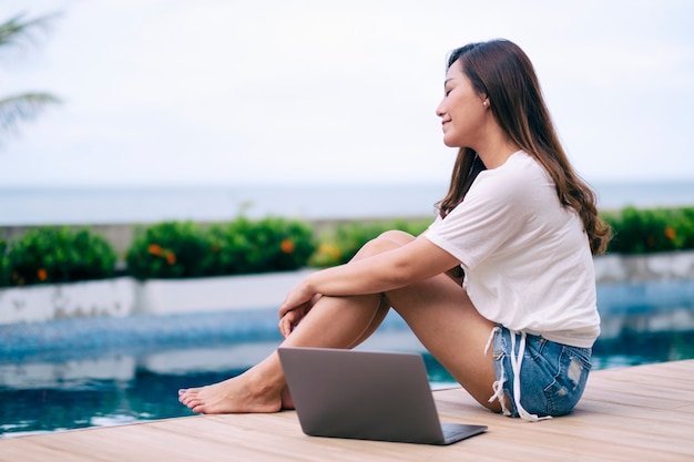 Una bella donna asiatica seduta a bordo piscina con un computer portatile