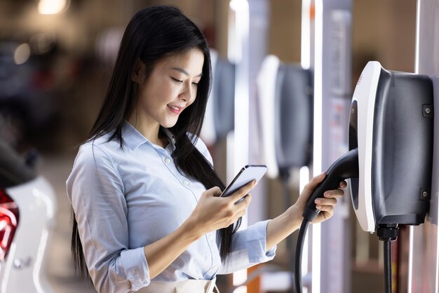 Beautiful asian woman standing at charging station in parking lot shopping mall Asian woman charging