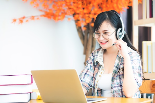 A beautiful Asian woman in Thailand wearing glasses and a plaid shirt, she is looking at a laptop computer and has headphones on the table. In the concept of learning or teleconferencing via online.