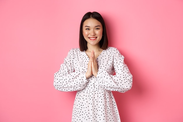 Beautiful asian woman thanking you, holding hands together in appreciation gesture, smiling happy at camera, standing grateful over pink.