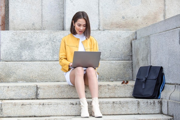 Красивая азиатка с ноутбуком Улыбающаяся азиатка сидит на лестнице и с ноутбуком Азиатская деловая женщина с ноутбуком