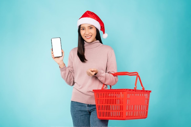 Bella donna asiatica che indossa il cappello di natale con lo smartphone in mano mockup di schermo vuoto e carrello rosso su sfondo blu.