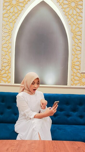Photo beautiful asian woman wearing a hijab candid expression of surprise pointing and checking her cell p