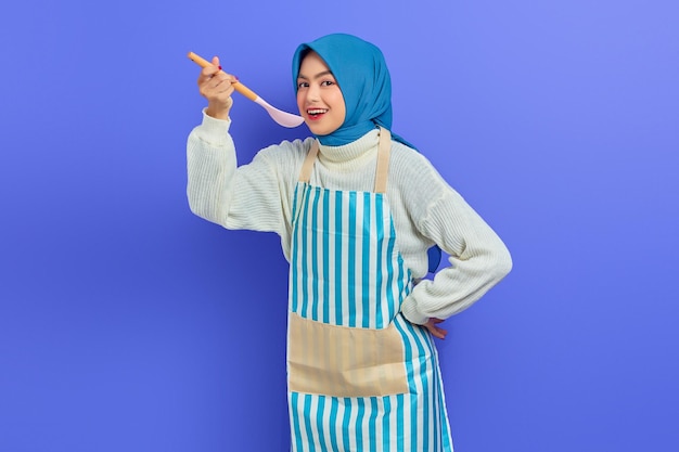 Beautiful Asian woman in white sweater covered in apron and hijab holding spoon, want to taste delicious food isolated over purple background. Housekeeping concept