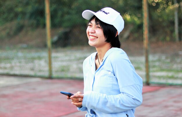 Bella donna asiatica con i capelli corti, indossa il cappello e sorride ampiamente sul campo da tennis