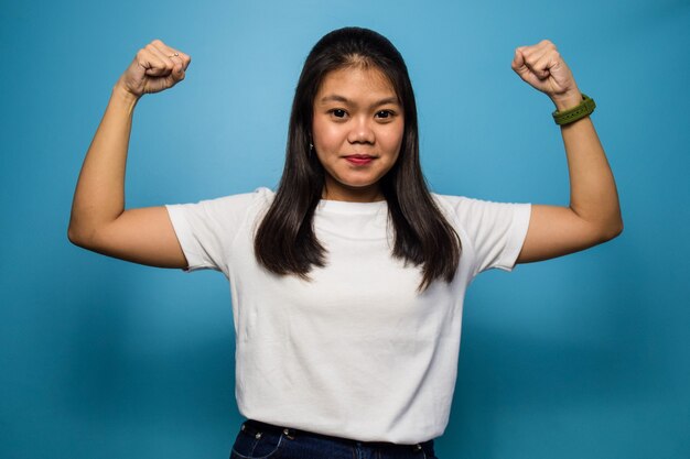 beautiful asian women using white Tshirt with blue isolated background raises arms and shows bicep