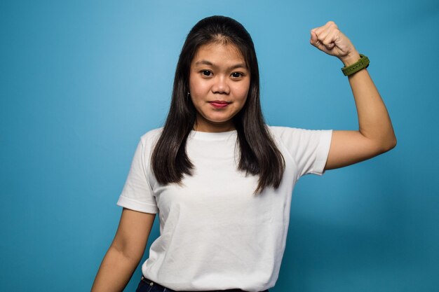 beautiful asian women using white Tshirt with blue isolated background raises arms and shows bicep