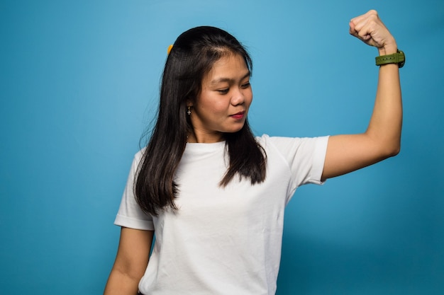 beautiful asian women using white Tshirt with blue isolated background raises arms and shows bicep