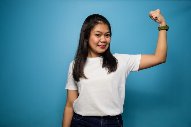 beautiful asian women using white Tshirt with blue isolated background raises arms and shows bicep