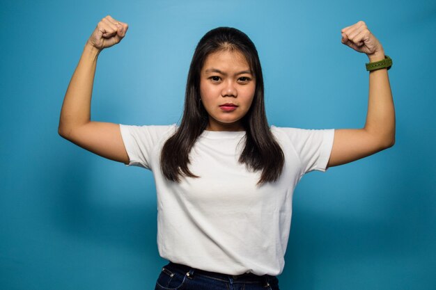 beautiful asian women using white Tshirt with blue isolated background raises arms and shows bicep