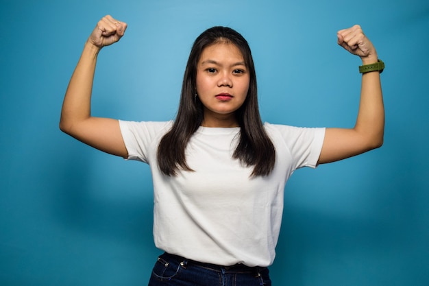 beautiful asian women using white Tshirt with blue isolated background raises arms and shows bicep